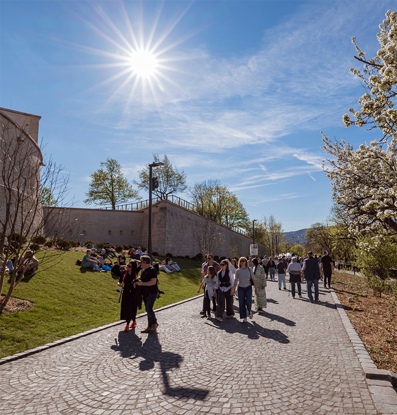 A Citadella megújult közparkjában látogatók sétálnak a kőburkolatú sétányon, míg mások a füves domboldalon pihennek a történelmi erődfalak mellett.
