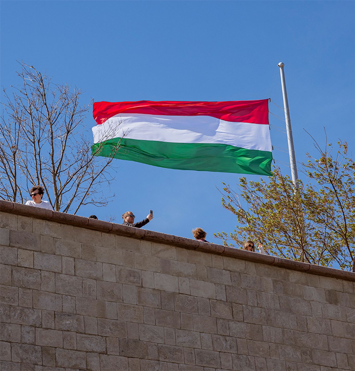 A Citadella területén magas zászlórúdon lobogó magyar nemzeti zászló, az előtérben a történelmi kőfalakkal és a sétányon tartózkodó látogatókkal.